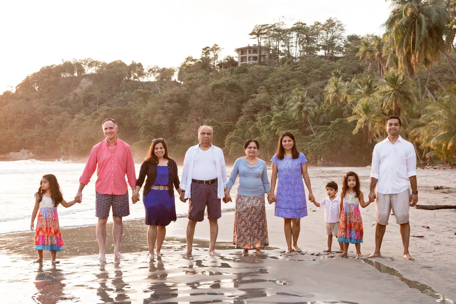 Three generations of one family standing together on a beach at sunset, holding hands.